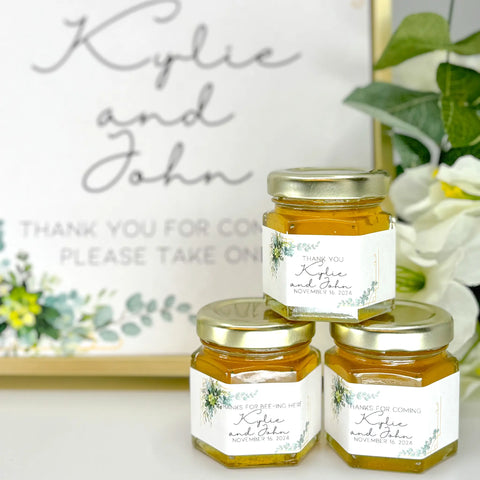 2 oz honey thank you wedding party shower favors on hexagon jars with gold lid and personalized labels. Labels feature gold frame with eucalyptus leaves with a variety of titles. Title shown include Thank You, Thanks for Bee-ing Here, and Thanks for Coming. Matching printable table sign in background.