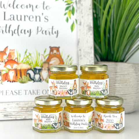 Favor table decor shown including honey birthday party favor jars topped with gold lids. Jars have labels featuring woodland animals and a variety of titles, including Our Little Birthday Girl, Our Little Birthday Boy, Thank You, Welcome to Lauren's Birthday Party, and Our Little Bear's Birthday. Table also shows a matching free printable sign and a plant.